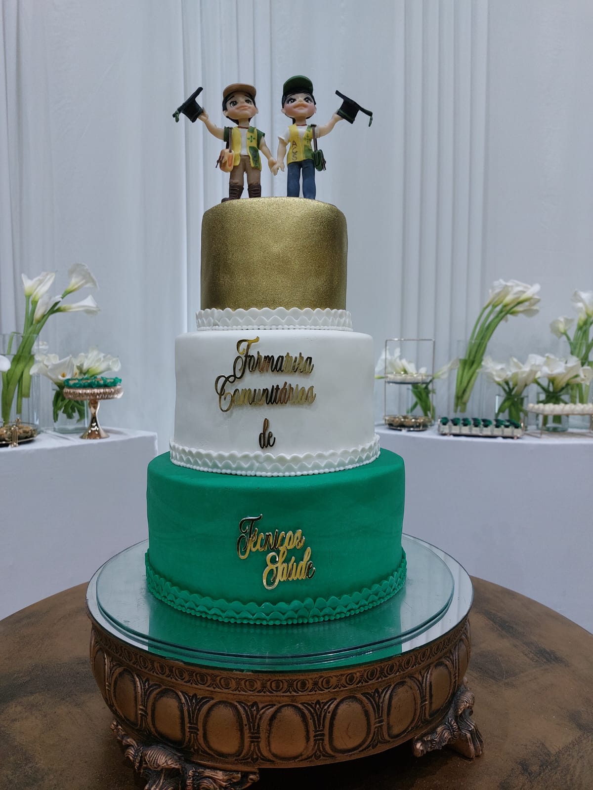 Bolo de formatura em verde, branco e dourado sobre suporte espelhado